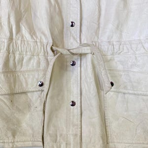 Puede incluir: Una chaqueta de cuero blanca con cintura de cord&oacute;n y cierre de botones. La chaqueta tiene dos bolsillos en la parte delantera y un cierre de bot&oacute;n plateado.