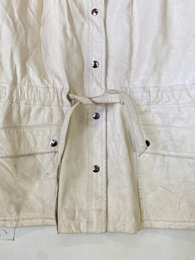 Puede incluir: Una chaqueta de cuero blanca con cintura de cord&oacute;n y cierre de botones. La chaqueta tiene dos bolsillos en la parte delantera y un cierre de bot&oacute;n plateado.