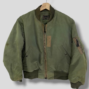 May include: Olive green bomber jacket with a brown zipper, pockets, and ribbed cuffs. The jacket has a brown patch on the chest and a small pocket on the left sleeve. It is hanging on a wooden hanger.