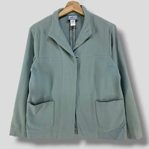 May include: A pale teal blazer with two front pockets and a notched collar hangs on a wooden hanger. The jacket has a single-button closure and a tailored fit. The fabric appears to be a soft, woven material.