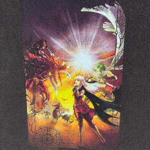 May include: Black t-shirt with a colourful fantasy illustration. The artwork shows a battle scene with a central female figure in red and black clothing, surrounded by other characters and a dragon. The background is a mix of orange and yellow hues.