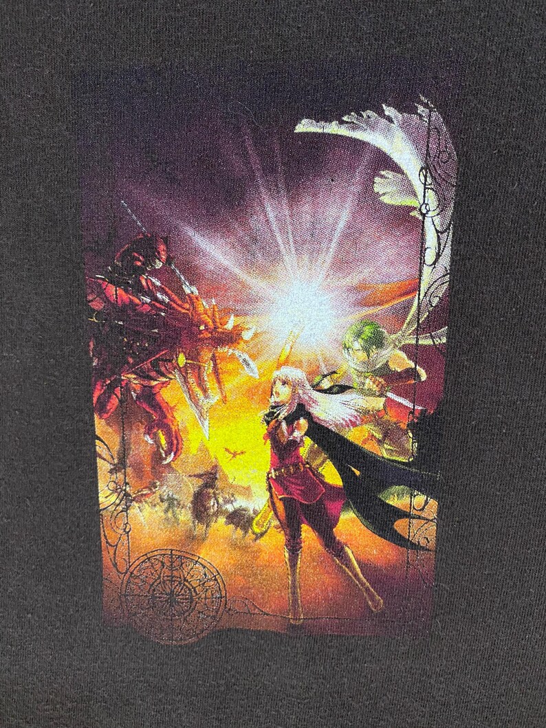 May include: Black t-shirt with a colourful fantasy illustration. The artwork shows a battle scene with a central female figure in red and black clothing, surrounded by other characters and a dragon. The background is a mix of orange and yellow hues.