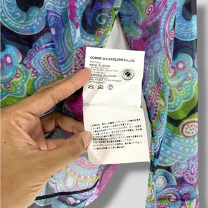 Puede incluir: Top transparente de manga larga con un vibrante estampado de cachemira en tonos azules, morados y verdes. La etiqueta dice "COMME des GAR&Ccedil;ONS Co., Ltd. MADE IN JAPAN". Hecho de 100% poli&eacute;ster.