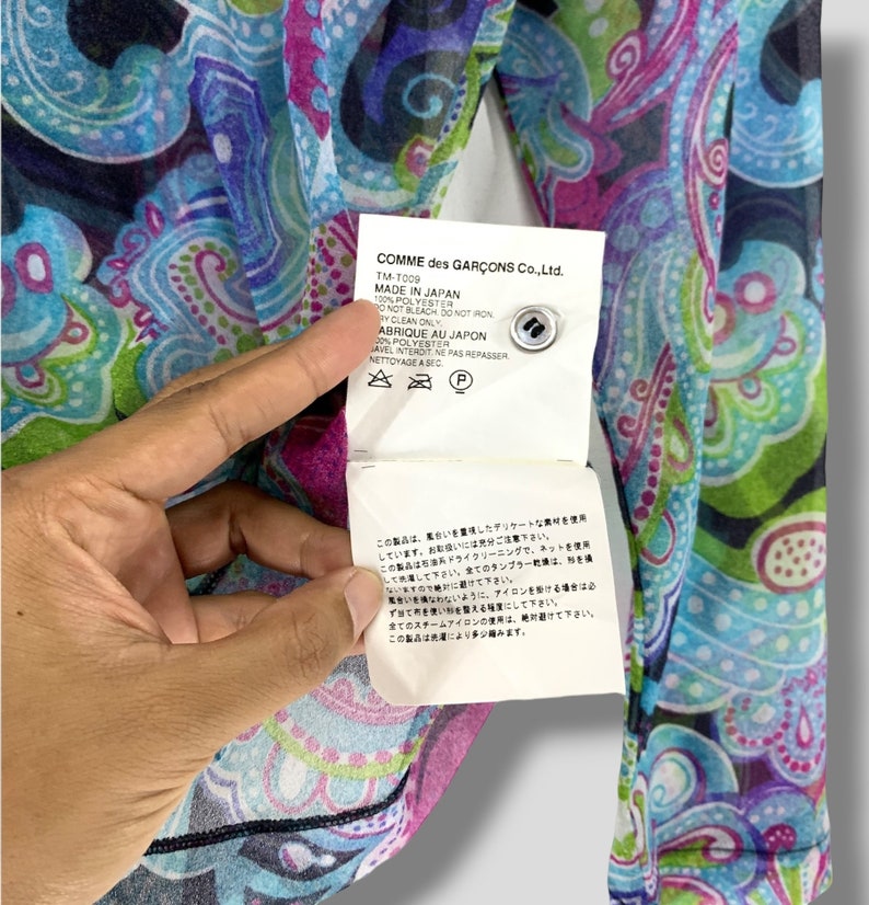 Puede incluir: Top transparente de manga larga con un vibrante estampado de cachemira en tonos azules, morados y verdes. La etiqueta dice "COMME des GAR&Ccedil;ONS Co., Ltd. MADE IN JAPAN". Hecho de 100% poli&eacute;ster.