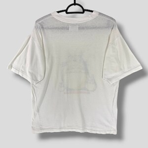 K&ouml;nnte beinhalten: Wei&szlig;es T-Shirt mit einer Grafik einer Zeichentrickfigur. Das T-Shirt h&auml;ngt an einem schwarzen Kleiderb&uuml;gel. Die Grafik hat gelbe, rote und schwarze Farben. Das T-Shirt hat kurze &Auml;rmel und einen Rundhalsausschnitt.