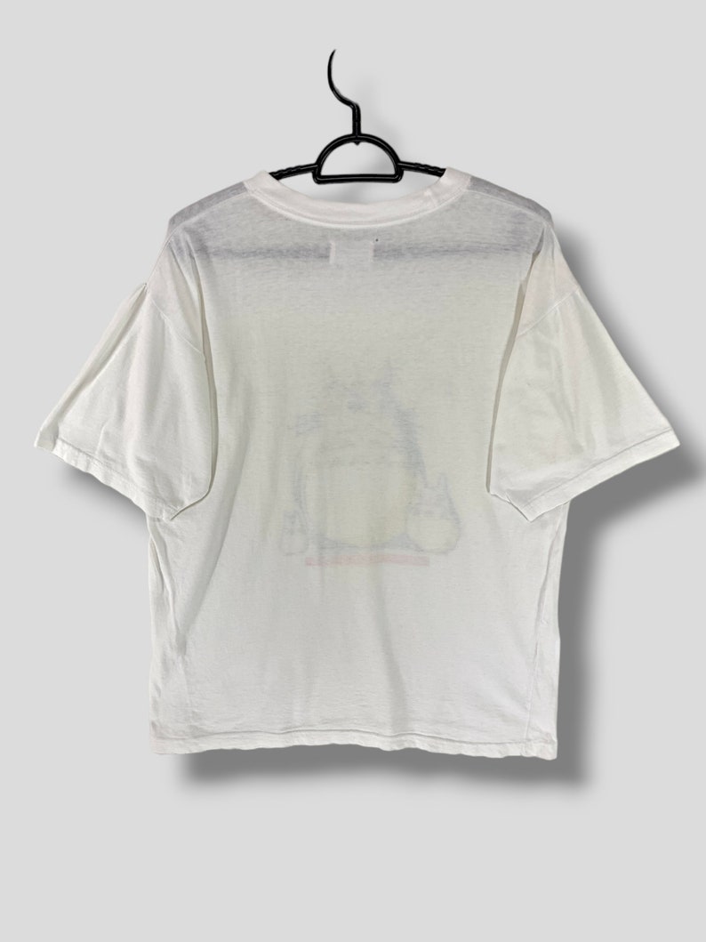 K&ouml;nnte beinhalten: Wei&szlig;es T-Shirt mit einer Grafik einer Zeichentrickfigur. Das T-Shirt h&auml;ngt an einem schwarzen Kleiderb&uuml;gel. Die Grafik hat gelbe, rote und schwarze Farben. Das T-Shirt hat kurze &Auml;rmel und einen Rundhalsausschnitt.