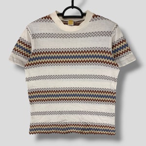 Può includere: T-shirt bianco a maniche corte con motivo a zig-zag marrone, blu e beige. La maglietta è appesa a un'appendiabiti nero. La parte superiore ha uno scollo tondo e una vestibilità leggermente corta.