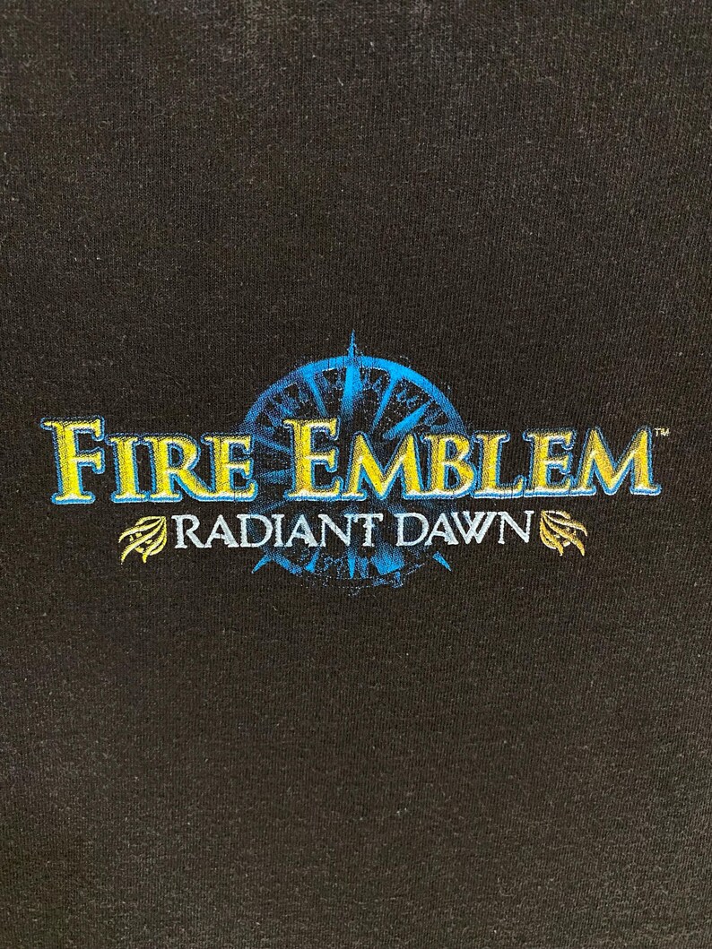 May include: Black fabric featuring the text "Fire Emblem Radiant Dawn" in gold and blue. The design includes a compass rose and decorative elements, suggesting a video game or fantasy theme. The text is stylized.