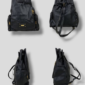 May include: Black leather backpack with gold-toned hardware, including zippers and a small rectangular logo plate. The backpack has a drawstring closure, two side pockets, and adjustable shoulder straps. White stitching accents the edges.