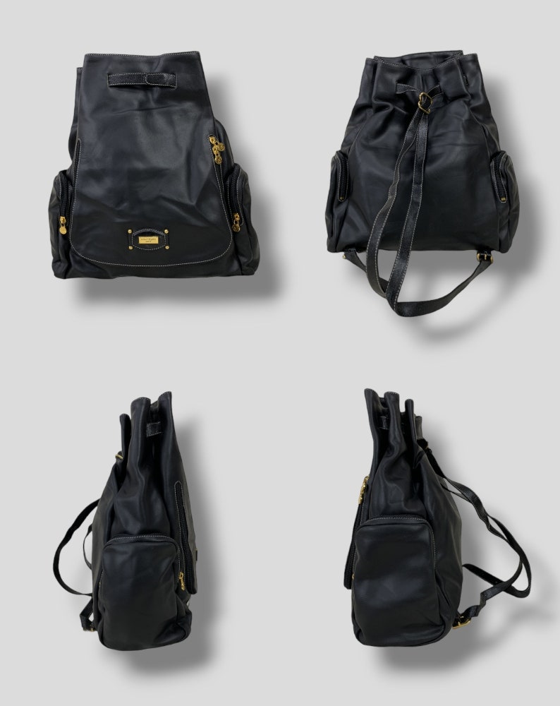May include: Black leather backpack with gold-toned hardware, including zippers and a small rectangular logo plate. The backpack has a drawstring closure, two side pockets, and adjustable shoulder straps. White stitching accents the edges.