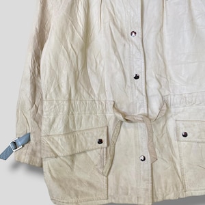 Puede incluir: Una chaqueta de cuero de color crema con cuello azul y cintur&oacute;n. La chaqueta tiene cierre de cremallera y dos bolsillos en la parte delantera. La chaqueta tiene un logotipo en el lado derecho del pecho.