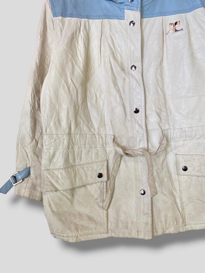 Puede incluir: Una chaqueta de cuero de color crema con cuello azul y cintur&oacute;n. La chaqueta tiene cierre de cremallera y dos bolsillos en la parte delantera. La chaqueta tiene un logotipo en el lado derecho del pecho.