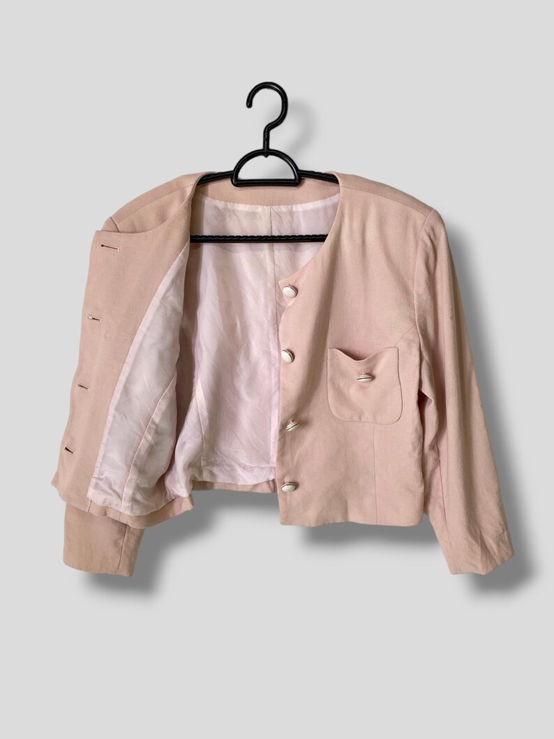 May include: A pale pink blazer with long sleeves and a button closure. The blazer has a single pocket on the right side and a white lining.