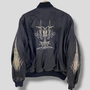 Puede incluir: Chaqueta bomber negra con diseños de pinstripes dorados en la espalda y las mangas. La chaqueta tiene cuello, puños y dobladillo de canalé negros. Los pinstripes son intrincados y estilizados.