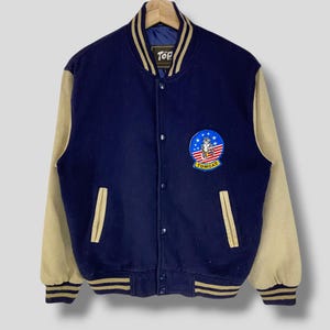 Puede incluir: Una chaqueta de béisbol azul marino y beige con un parche de logotipo de Top Cat en el pecho izquierdo. La chaqueta tiene cierre de presión y ribetes de rayas en los puños y el dobladillo.