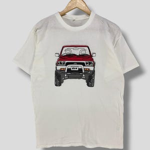 Puede incluir: Camiseta blanca con una ilustración roja y negra de un vehículo 4x4. El vehículo tiene una carrocería roja, neumáticos negros y el texto "RV4" en la parte delantera. La camiseta está colgada en una percha de madera.