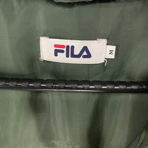 May include: Close-up of a green jacket interior with a white label. The label features the brand name "FILA" in red and navy blue, with the letter "M" to the right. The jacket is hanging on a black hanger.