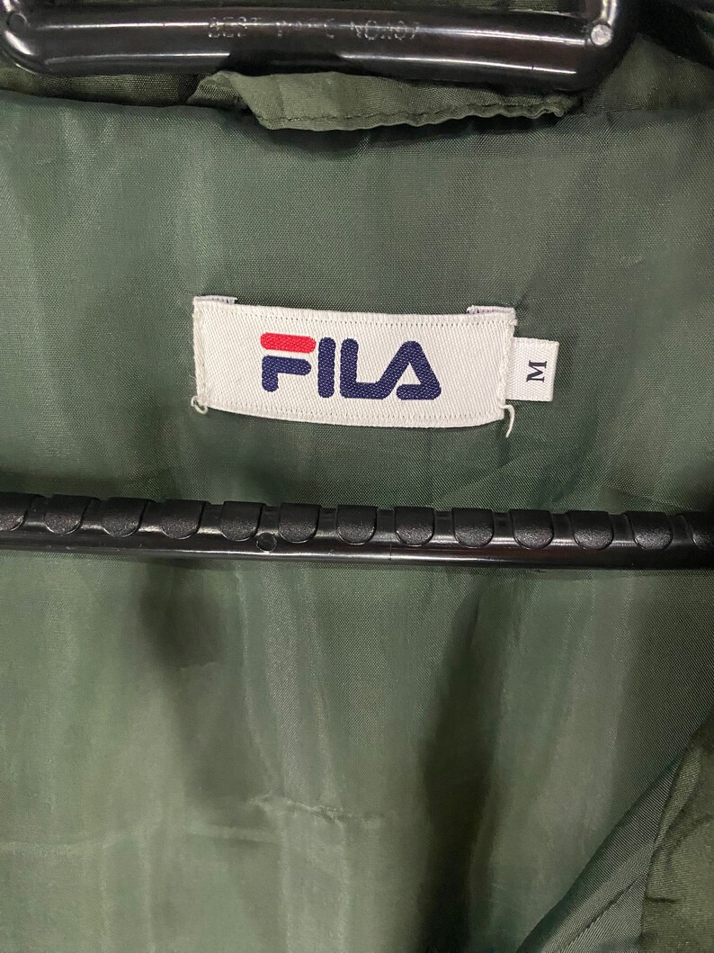 May include: Close-up of a green jacket interior with a white label. The label features the brand name "FILA" in red and navy blue, with the letter "M" to the right. The jacket is hanging on a black hanger.