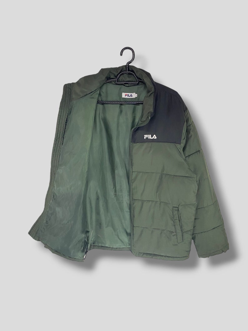 May include: Olive green puffer jacket with a black panel on the upper chest and collar. The jacket features the "FILA" logo in white on the chest. The jacket is hanging on a black hanger.
