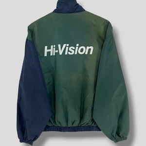 Może przedstawiać: Zielona i granatowa kurtka wiatrówka z białym napisem "Hi-Vision" na plecach. Kurtka ma wysoki kołnierz oraz prążkowany dół i mankiety. Rękawy są granatowe, a korpus zielony.