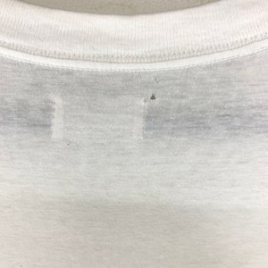 K&ouml;nnte beinhalten: Nahaufnahme des Ausschnitts eines wei&szlig;en T-Shirts. Das Shirt hat einen Rundhalsausschnitt und scheint aus einem d&uuml;nnen, leichten Stoff zu bestehen. Es gibt kleine L&ouml;cher im Stoff, die auf Abnutzung hindeuten.