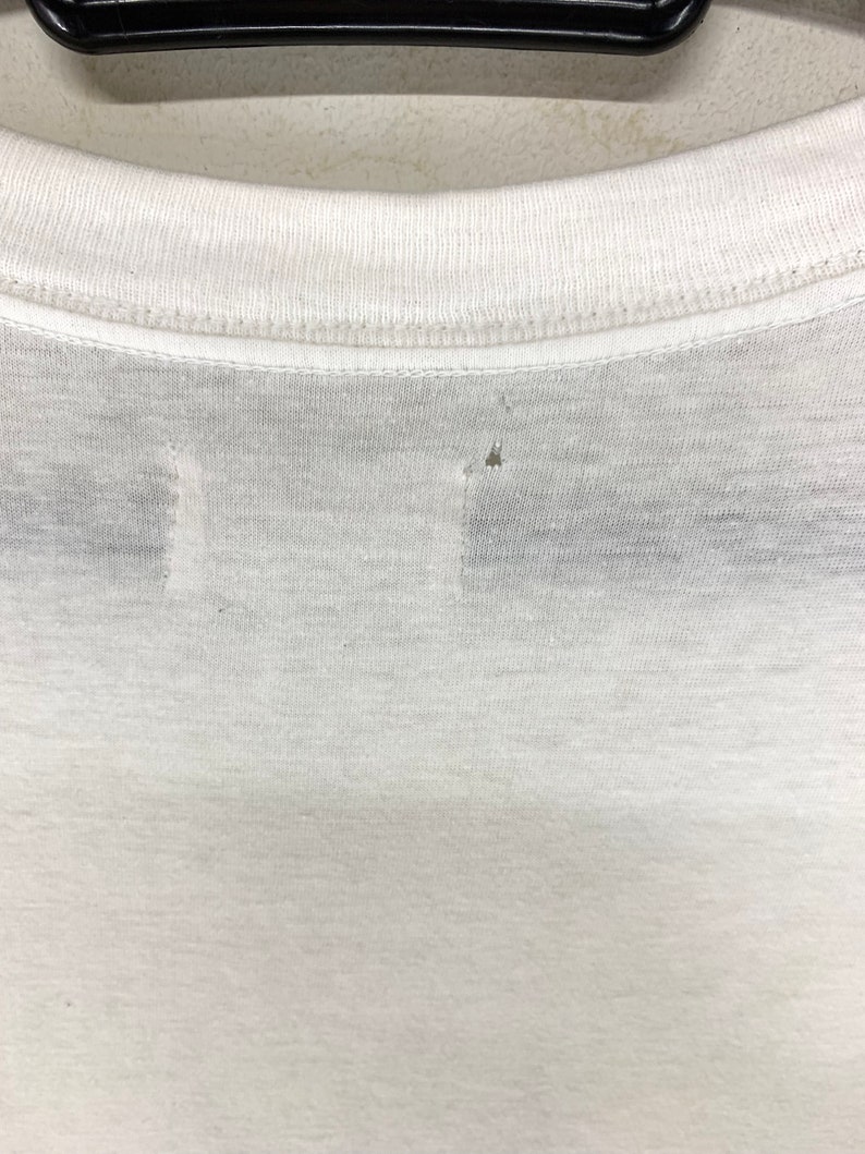 K&ouml;nnte beinhalten: Nahaufnahme des Ausschnitts eines wei&szlig;en T-Shirts. Das Shirt hat einen Rundhalsausschnitt und scheint aus einem d&uuml;nnen, leichten Stoff zu bestehen. Es gibt kleine L&ouml;cher im Stoff, die auf Abnutzung hindeuten.