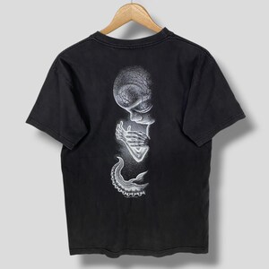 Puede incluir: Una camiseta negra de manga corta con un estampado gráfico blanco de puntos en la espalda. El diseño muestra una figura estilizada de tipo alienígena con una cabeza grande, manos delgadas cerca de la cara y un apéndice tentacular abajo.