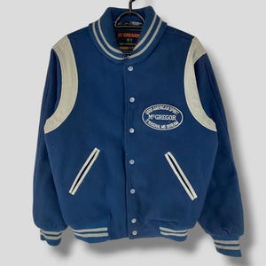 May include: A blue varsity jacket with white and black stripes on the sleeves and collar. The jacket has a white leather patch on the left side with the text "GOOD AMERICAN SPIRIT McGREGOR ORIGINAL MD DREAM".