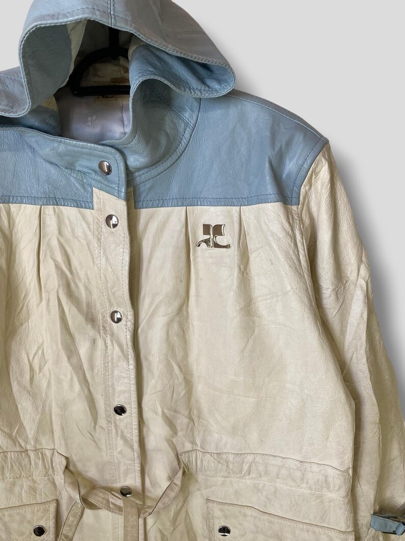 Puede incluir: Una chaqueta de cuero blanca y azul claro con capucha. La chaqueta tiene cierre de botones y cintur&oacute;n. La chaqueta tiene un logotipo en la parte delantera que dice "JC".