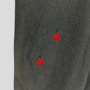 May include: Close-up of a dark gray fabric with two small holes. The fabric has a slightly textured appearance and is evenly lit, highlighting the details of the material. The holes are indicated by red arrows.