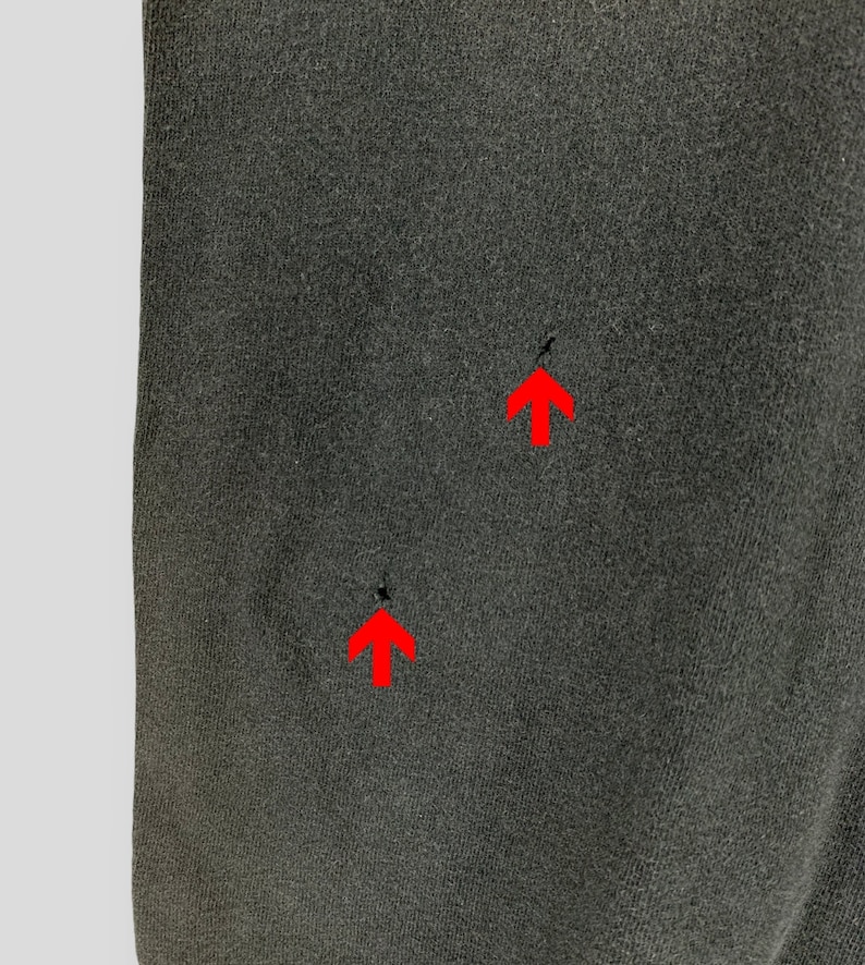 May include: Close-up of a dark gray fabric with two small holes. The fabric has a slightly textured appearance and is evenly lit, highlighting the details of the material. The holes are indicated by red arrows.