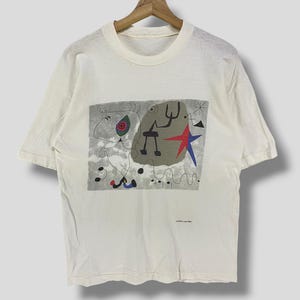 Puede incluir: Camiseta de color crema con un dise&ntilde;o abstracto colorido. El gr&aacute;fico incluye una estrella roja, formas azules y figuras negras sobre un fondo gris. La camiseta est&aacute; colgada en una percha de madera.