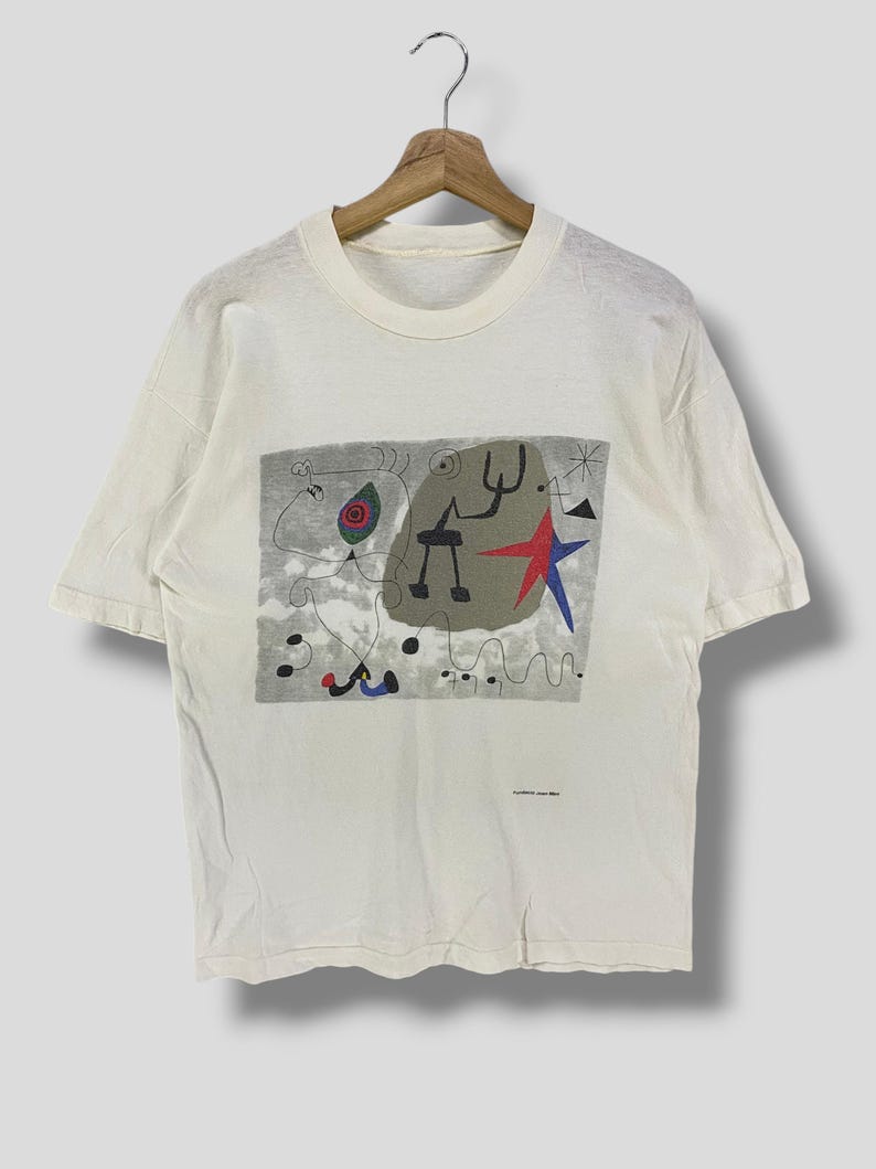 Puede incluir: Camiseta de color crema con un dise&ntilde;o abstracto colorido. El gr&aacute;fico incluye una estrella roja, formas azules y figuras negras sobre un fondo gris. La camiseta est&aacute; colgada en una percha de madera.