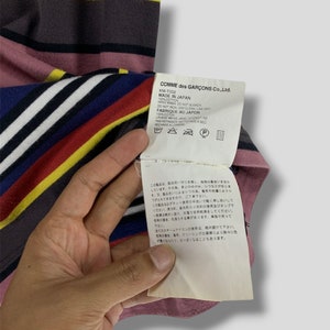 May include: A striped t-shirt with a tag from Comme des Gar&ccedil;ons. The shirt features horizontal stripes in various colors, including pink, yellow, blue, and white. The tag provides care instructions and states "Made in Japan."