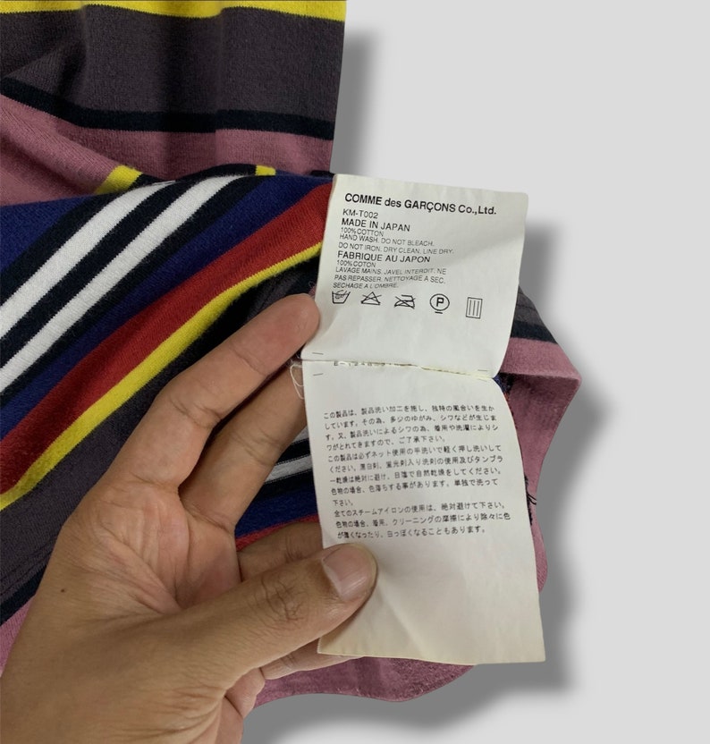 May include: A striped t-shirt with a tag from Comme des Gar&ccedil;ons. The shirt features horizontal stripes in various colors, including pink, yellow, blue, and white. The tag provides care instructions and states "Made in Japan."