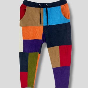 Puede incluir: Pantalones deportivos de patchwork multicolor con cintura con cordón. Los pantalones presentan una variedad de colores, incluyendo azul, naranja, marrón, rojo y morado. El diseño está formado por parches rectangulares de tela.