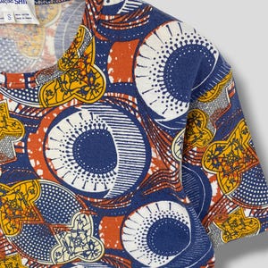 K&ouml;nnte beinhalten: Ein kurz&auml;rmeliges T-Shirt mit einem lebendigen Muster in Blau-, Orange-, Gelb- und Wei&szlig;t&ouml;nen. Das Design zeigt gro&szlig;e kreisf&ouml;rmige und geometrische Formen. Das Shirt h&auml;ngt an einem Holzb&uuml;gel.