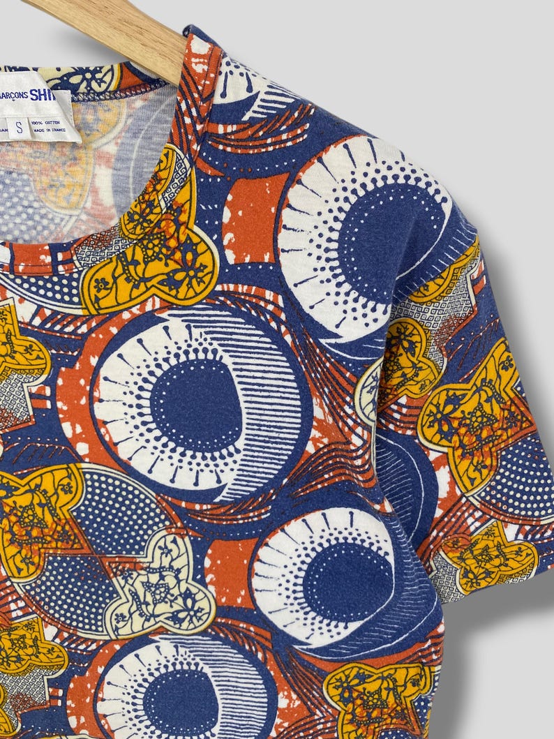 K&ouml;nnte beinhalten: Ein kurz&auml;rmeliges T-Shirt mit einem lebendigen Muster in Blau-, Orange-, Gelb- und Wei&szlig;t&ouml;nen. Das Design zeigt gro&szlig;e kreisf&ouml;rmige und geometrische Formen. Das Shirt h&auml;ngt an einem Holzb&uuml;gel.