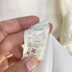 K&ouml;nnte beinhalten: Nahaufnahme eines wei&szlig;en Stoffetiketts mit schwarzem Text. Das Etikett enth&auml;lt den Text "ATN-1849", Stoffprozents&auml;tze und Pflegehinweise. Das Etikett wird von einer Hand gehalten.