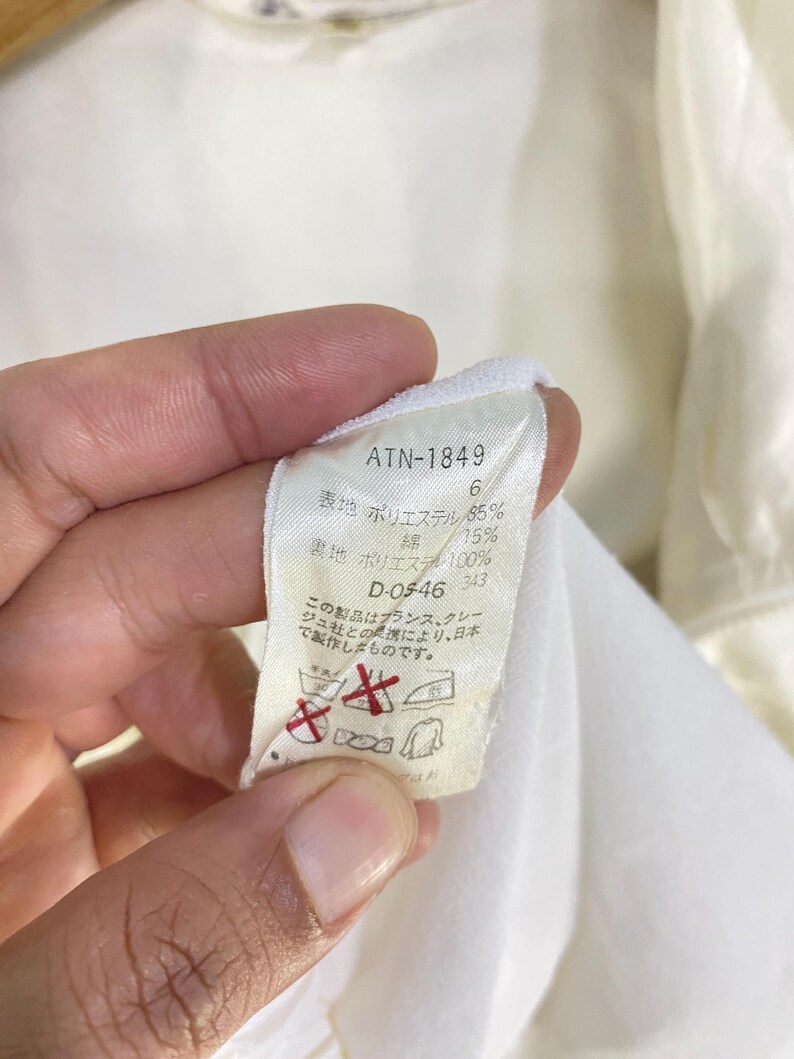 K&ouml;nnte beinhalten: Nahaufnahme eines wei&szlig;en Stoffetiketts mit schwarzem Text. Das Etikett enth&auml;lt den Text "ATN-1849", Stoffprozents&auml;tze und Pflegehinweise. Das Etikett wird von einer Hand gehalten.