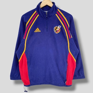 May include: A navy blue quarter-zip fleece pullover featuring the Adidas logo on the right chest and the Real Federación Española de Fútbol crest on the left. The garment includes red and yellow stripes along the shoulders and red side panels.