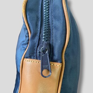 May include: Close-up of a navy blue bag with a brown leather accent and a matching navy blue zipper. The zipper is partially open, revealing the interior. The bag has a rectangular shape.