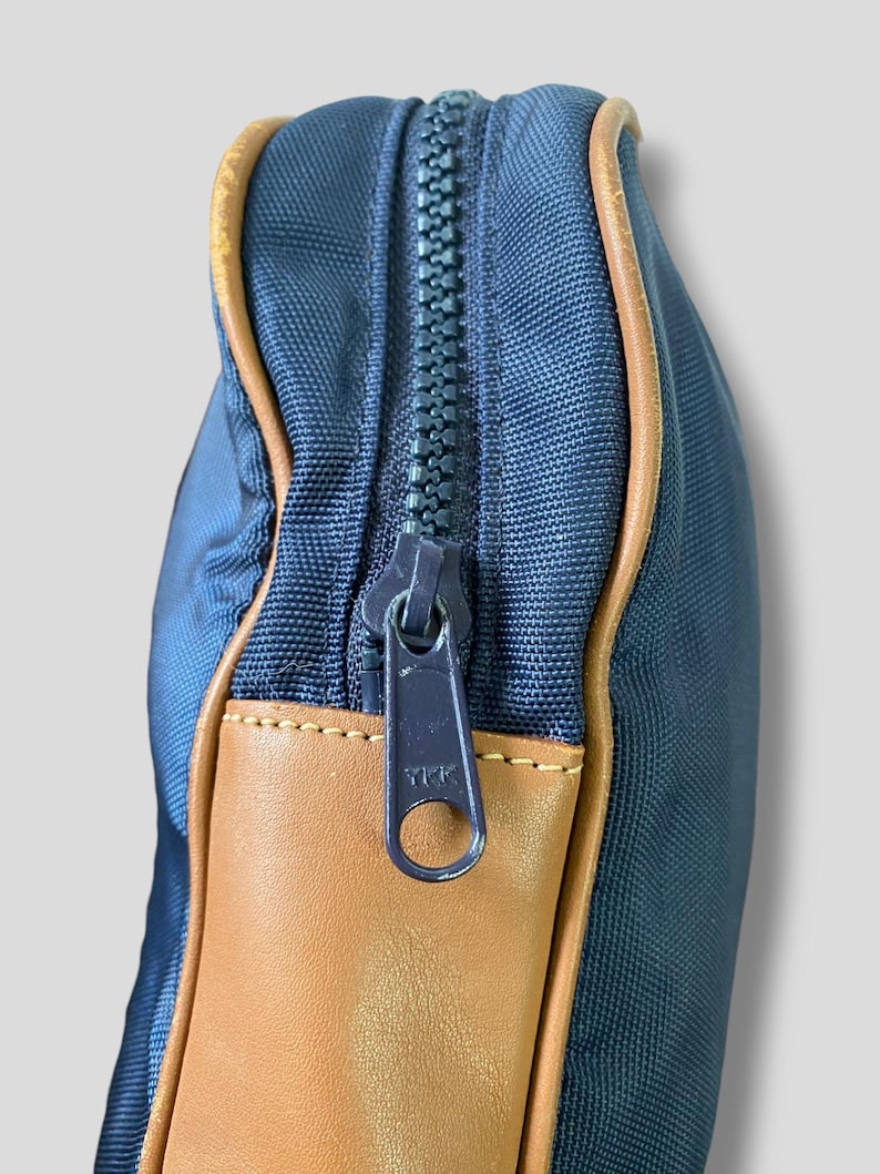 May include: Close-up of a navy blue bag with a brown leather accent and a matching navy blue zipper. The zipper is partially open, revealing the interior. The bag has a rectangular shape.