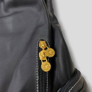 May include: Close-up of a black leather bag with gold-colored zippers and coin-shaped zipper pulls. The bag has white stitching and a textured surface. The zipper pulls have the text "Courreges Paris".