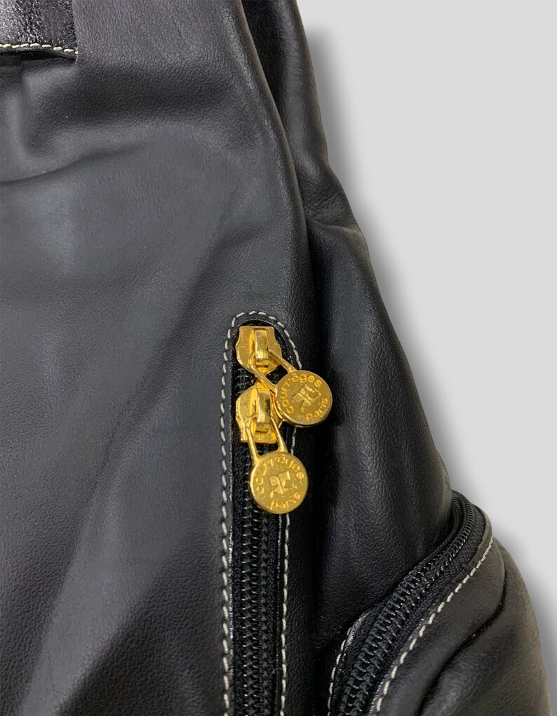 May include: Close-up of a black leather bag with gold-colored zippers and coin-shaped zipper pulls. The bag has white stitching and a textured surface. The zipper pulls have the text "Courreges Paris".