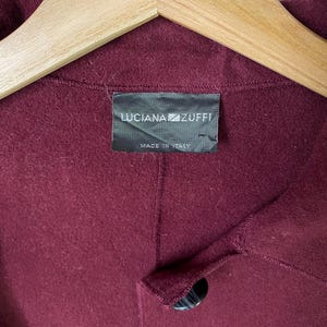 Puede incluir: Un abrigo de lana burdeos con un bot&oacute;n negro y una etiqueta que dice "LUCIANA ZUFFI MADE IN ITALY."