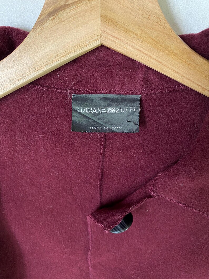 Puede incluir: Un abrigo de lana burdeos con un bot&oacute;n negro y una etiqueta que dice "LUCIANA ZUFFI MADE IN ITALY."