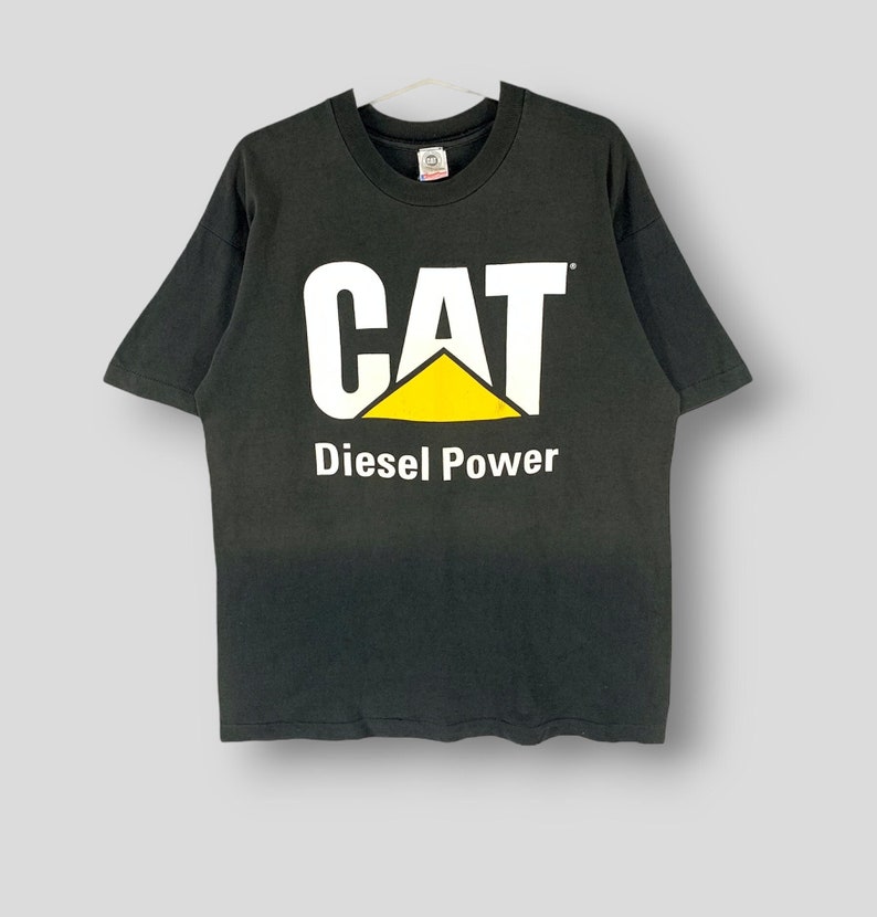 May include: Black t-shirt with the CAT logo in white and yellow, and the words "Diesel Power" in white below the logo. The shirt is hanging on a white hanger.