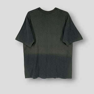 May include: A dark gray t-shirt with a faded, ombre effect. The shirt has short sleeves and a crew neck. The fabric appears to be soft and worn, giving it a vintage look. The shirt is displayed on a white background.