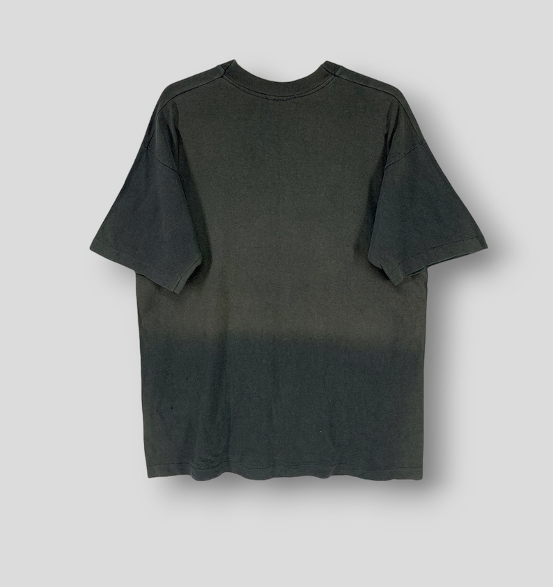 May include: A dark gray t-shirt with a faded, ombre effect. The shirt has short sleeves and a crew neck. The fabric appears to be soft and worn, giving it a vintage look. The shirt is displayed on a white background.